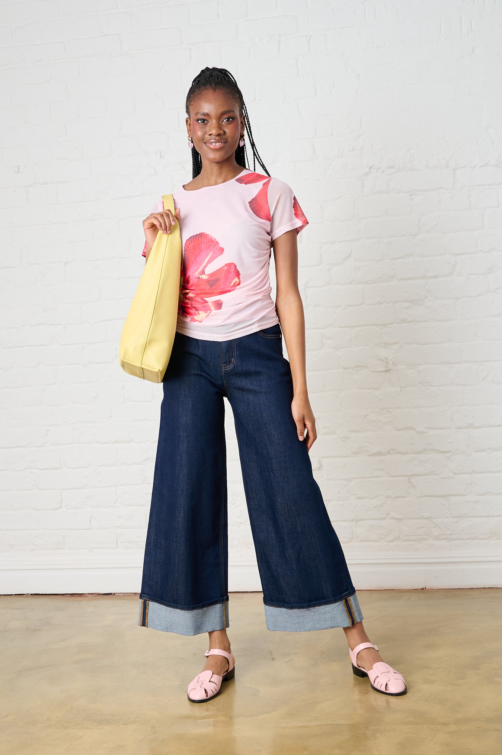 Woman wearing a pink floral top and blue wide-leg pants, holding a yellow bag against a white brick wall.