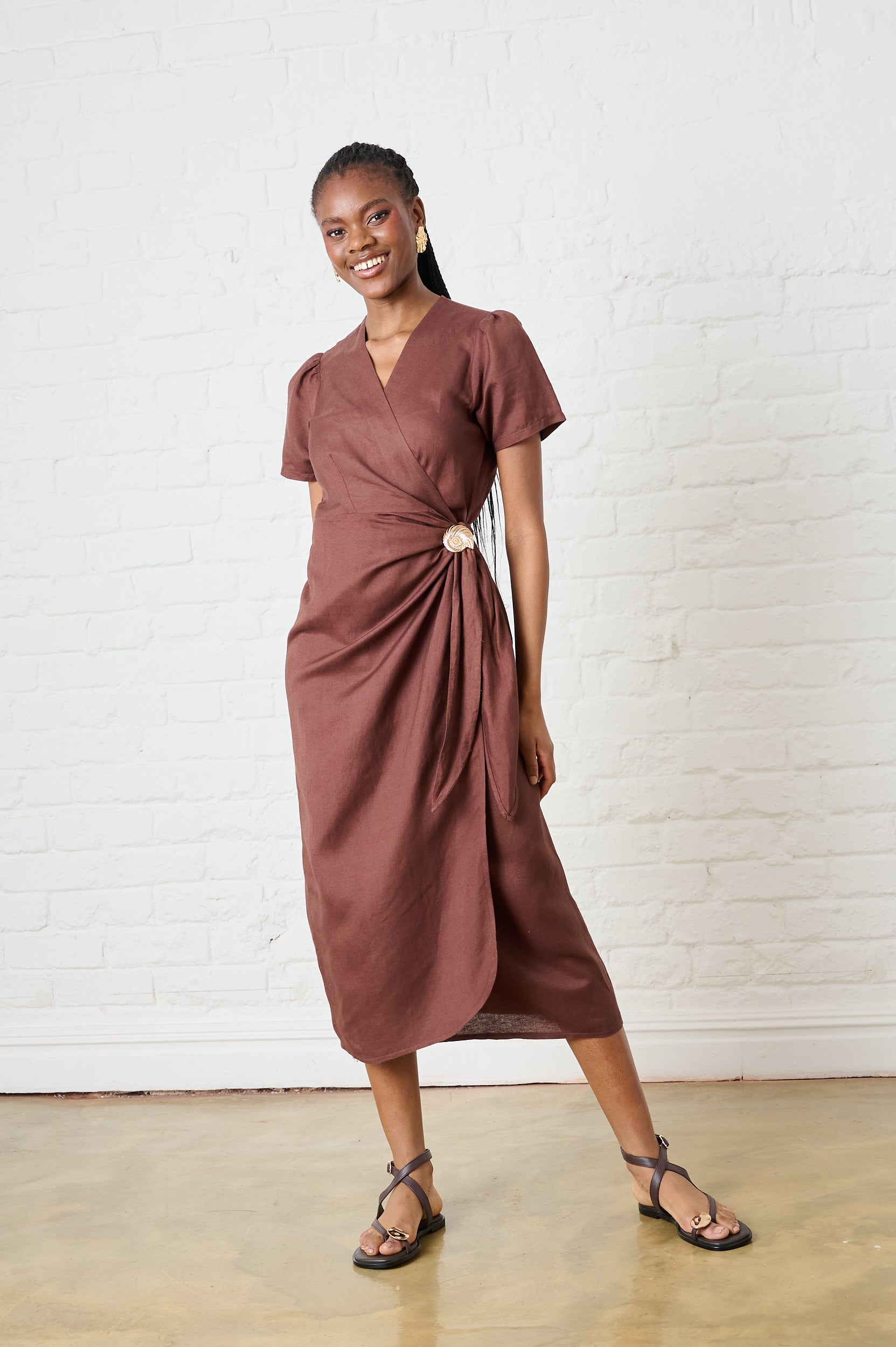Model is wearing a chocolate brown wrap dress with a gold shell detail and brown sandals