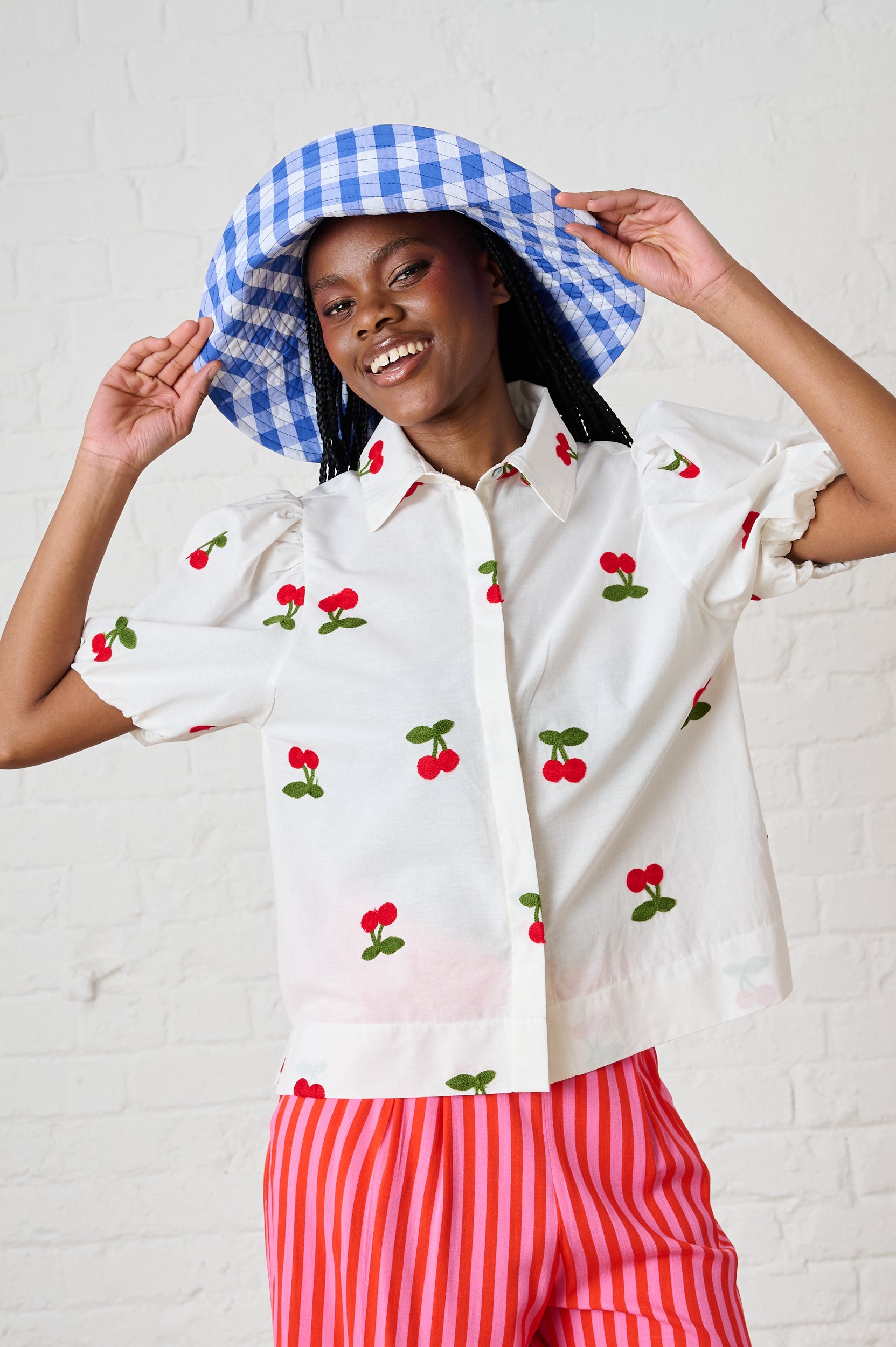 Woman wearing a white blouse with red cherry pattern and red striped pants, holding a blue and white checkered umbrella.