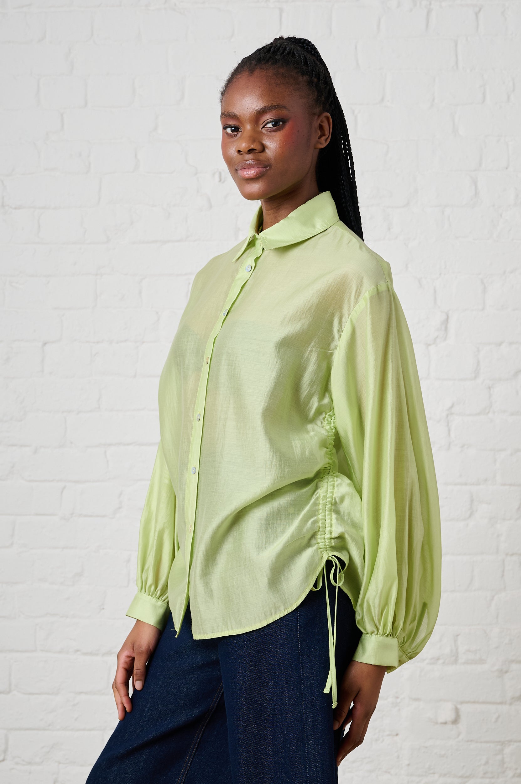 Woman wearing a light green blouse with long sleeves against a white brick wall.