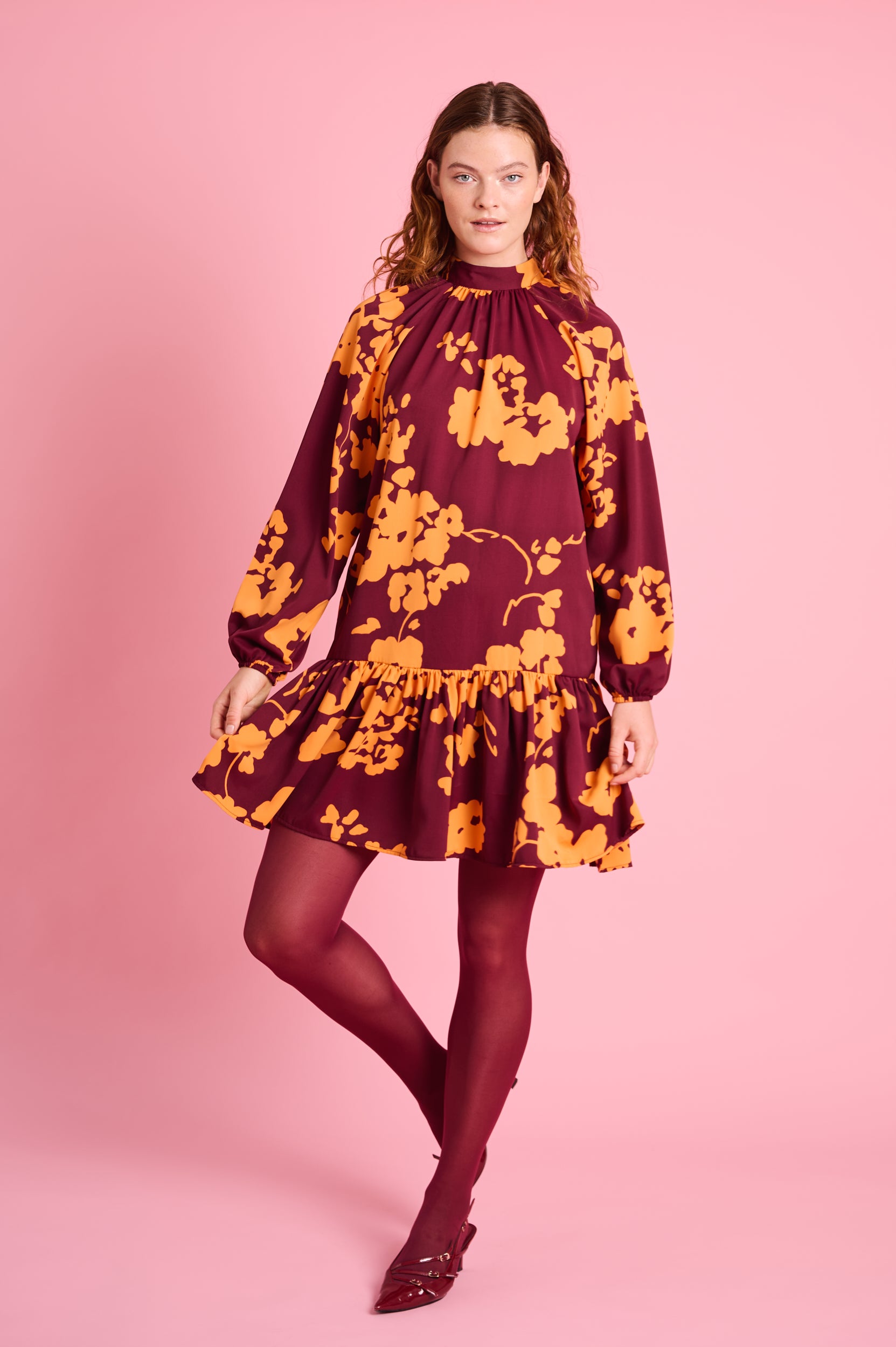 Model is wearing a long sleeve dress with orange flowers , burgundy tights and burgundy heels