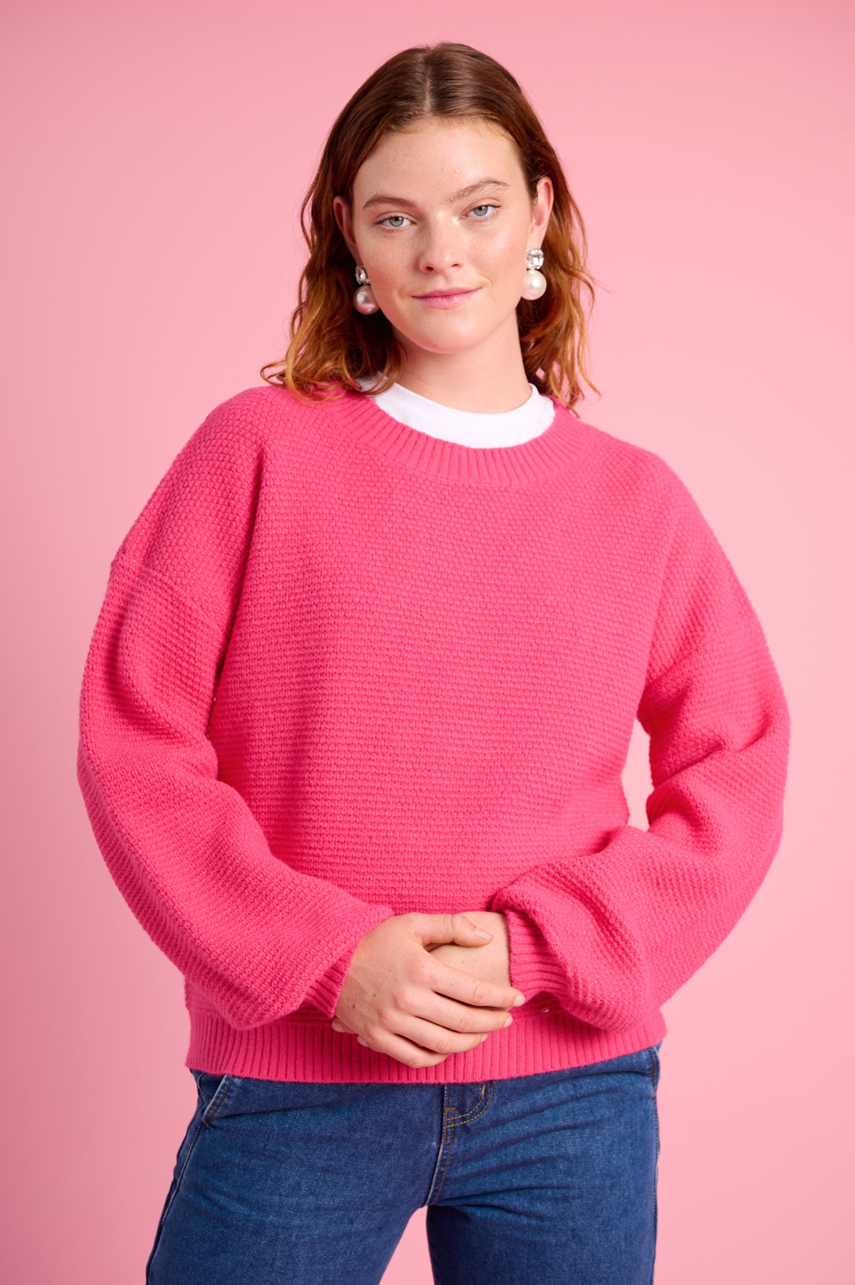 Model is wearing a pink jersey with blue jeans and pearl earrings