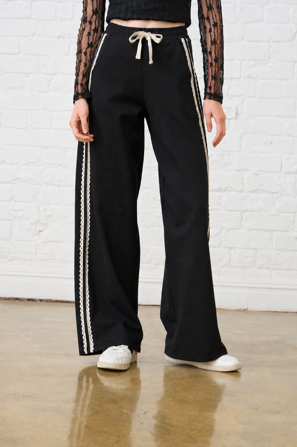 Model is wearing black wide leg pants with a lace trim along the side and white sneakers