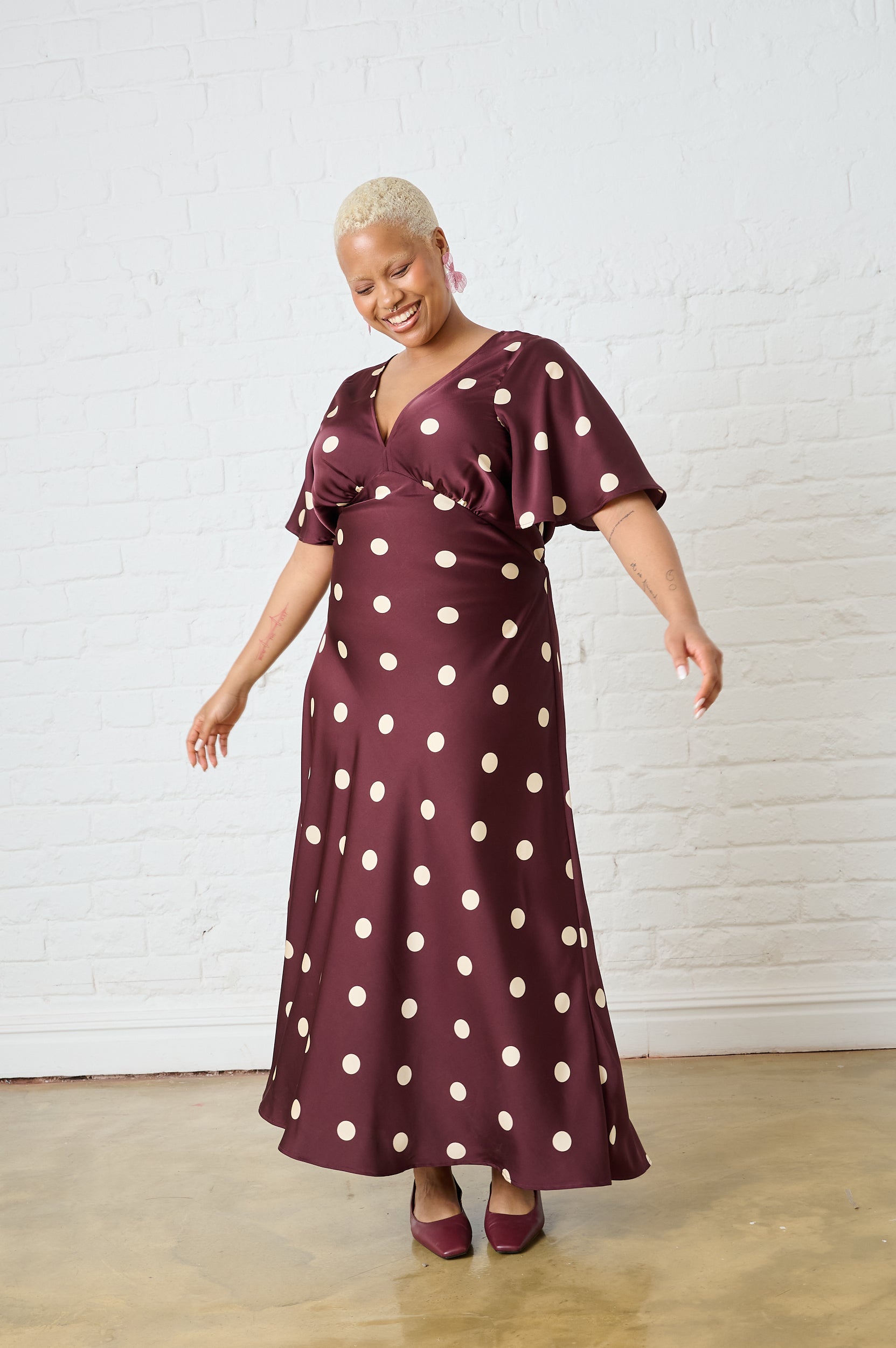 Woman wearing a burgundy dress with white polka dots against a white brick wall.