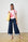 Woman wearing a pink floral top and blue wide-leg pants, holding a yellow bag against a white brick wall.