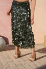 A model wearing a dark olive green sequined midi skirt and beige block-heel sandals, standing outdoors against a pink stucco wall.