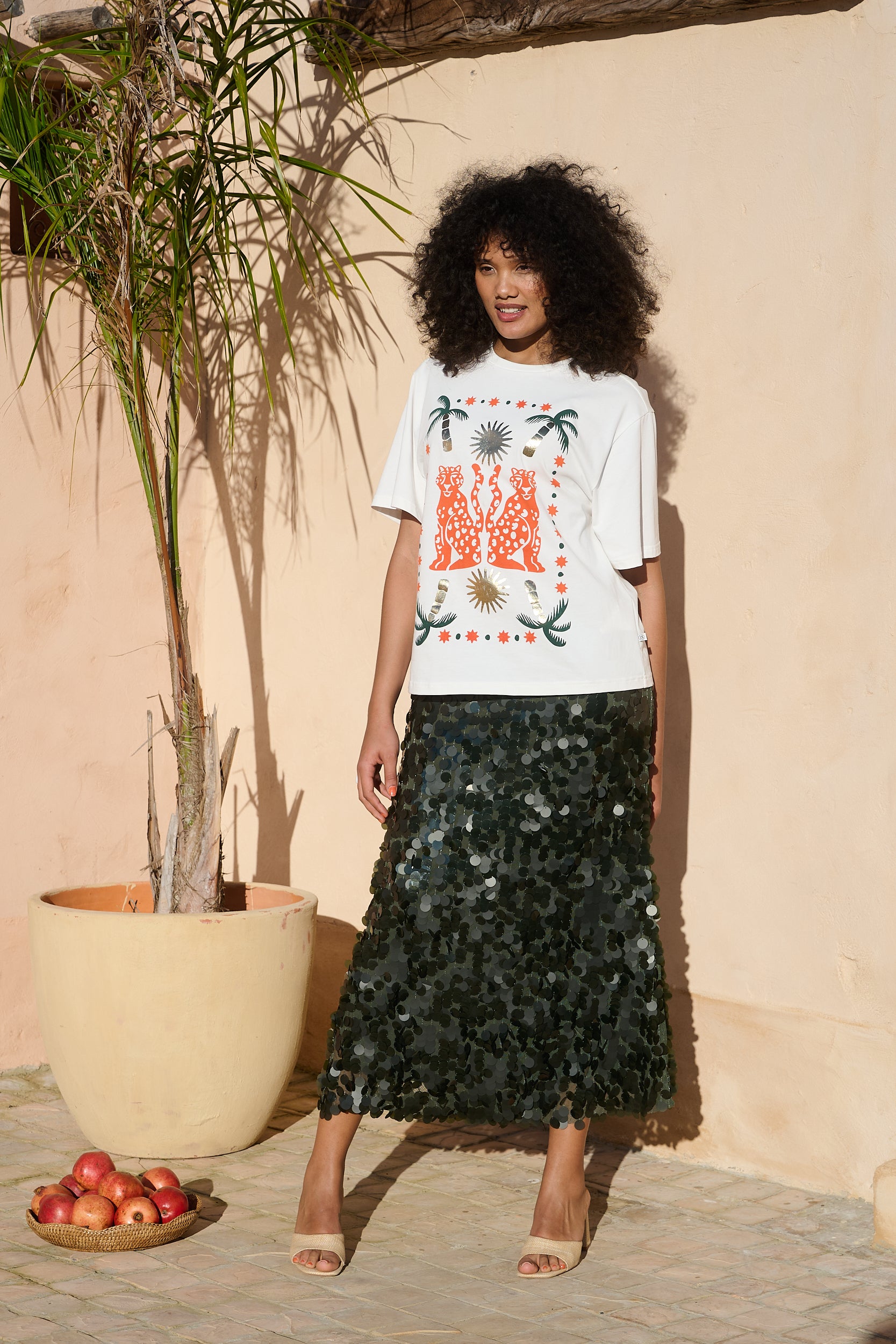 A model stands outdoors against a beige stucco wall wearing a white graphic T-shirt with orange and green print and a dark green sequined midi skirt. A potted palm and a small bowl of red apples are visible beside her.