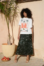 A model stands outdoors against a beige stucco wall wearing a white graphic T-shirt with orange and green print and a dark green sequined midi skirt. A potted palm and a small bowl of red apples are visible beside her.