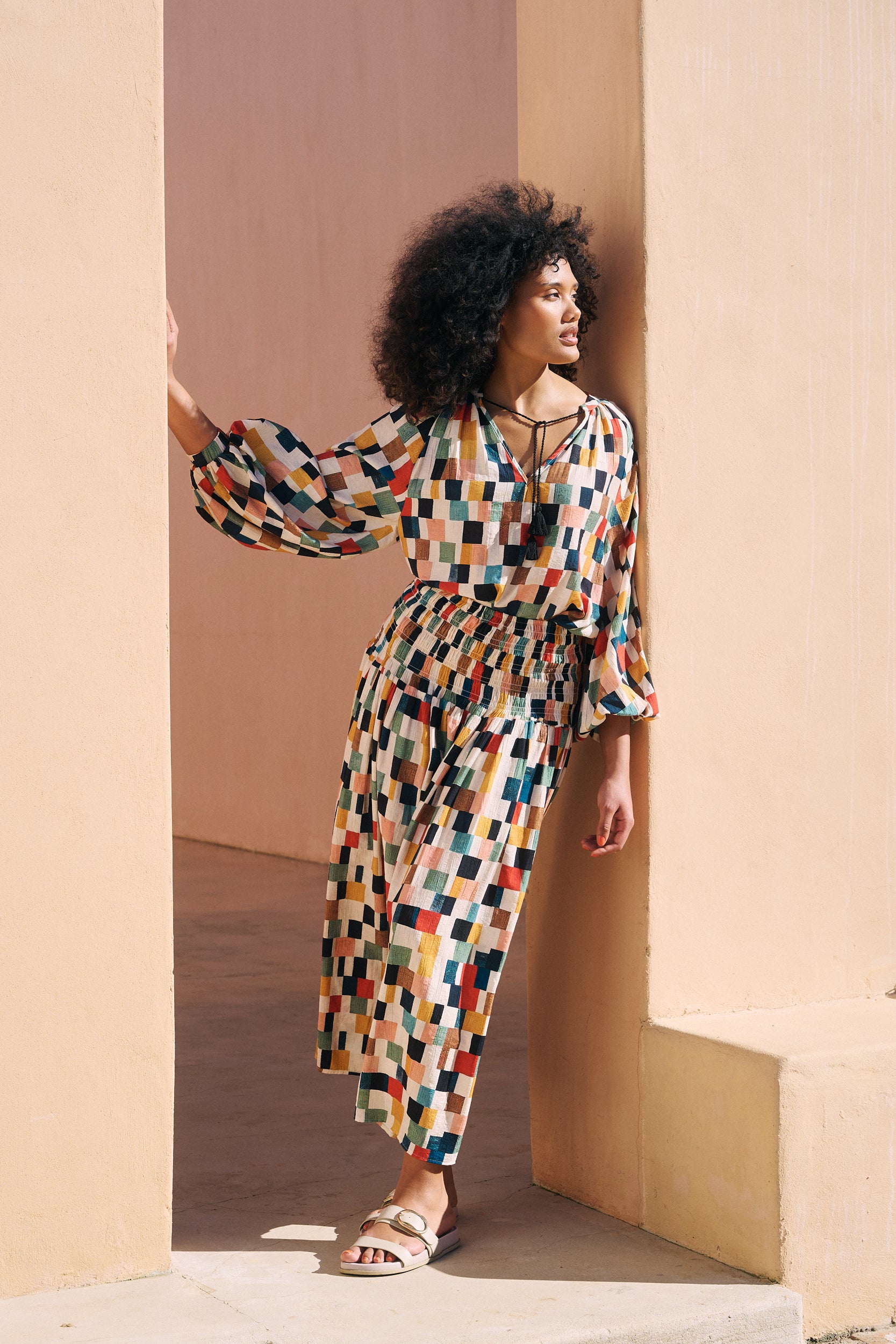 A woman with curly black hair models a long-sleeved maxi dress featuring a colorful geometric checkered pattern, leaning against a beige stucco wall in a doorway.