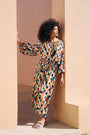 A woman with curly black hair models a long-sleeved maxi dress featuring a colorful geometric checkered pattern, leaning against a beige stucco wall in a doorway.