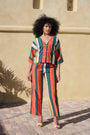 A model with curly black hair stands outdoors against a beige stucco wall wearing a vibrant, multicolored striped two-piece set consisting of a cropped V-neck top with front buttons and matching wide-leg cropped pants.