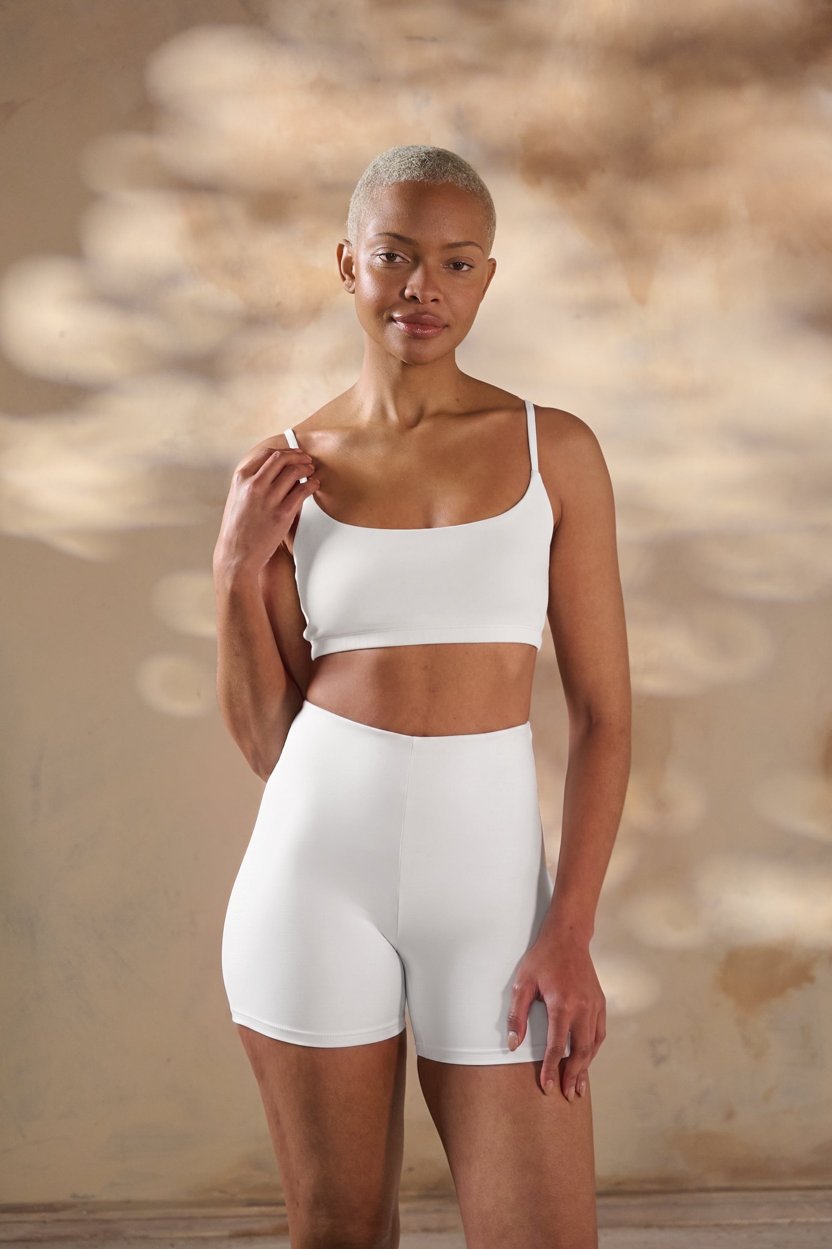 A woman modeling a white two-piece shapewear set consisting of a cropped bra and high-waisted shorts, set against a warm, textured beige wall with soft light shadows.
