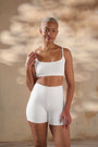 A woman modeling a white two-piece shapewear set consisting of a cropped bra and high-waisted shorts, set against a warm, textured beige wall with soft light shadows.