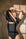 A model with short blonde hair wearing a black one-piece shapewear bodysuit, lifting the short sleeves to reveal a cutout at the chest, set against a textured beige wall with dramatic window shadows.