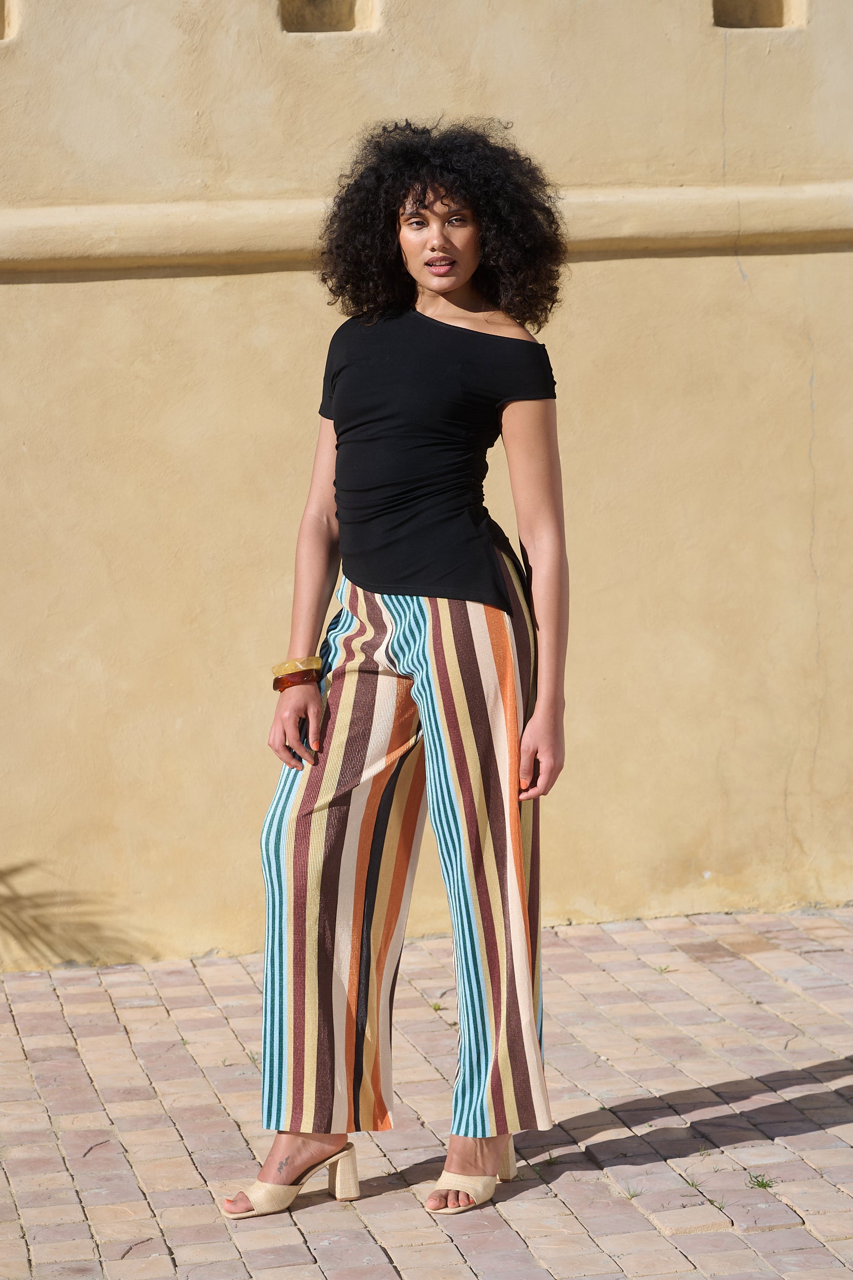 A woman stands outdoors against a beige stucco wall wearing a black off-the-shoulder top and wide-leg striped pants with vertical stripes in brown, orange, cream, and light blue. She is wearing beige heeled sandals.