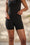 A model wearing a black shapewear top and matching black shorts, shown from the chest down against a softly blurred beige background.