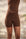 A person wearing fitted brown shapewear shorts with a matching brown top, shown from the chest down against a softly blurred beige background with a hint of greenery.
