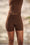 A person wearing fitted brown shapewear shorts with a matching brown top, shown from the chest down against a softly blurred beige background with a hint of greenery.