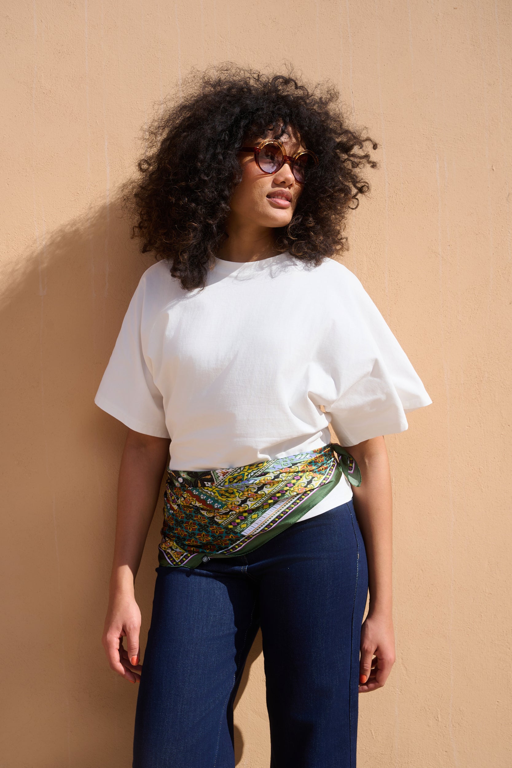 A woman with curly black hair and glasses wearing a white short-sleeve top, dark blue pants, and a green and yellow patterned scarf tied around her waist, standing against a beige wall.