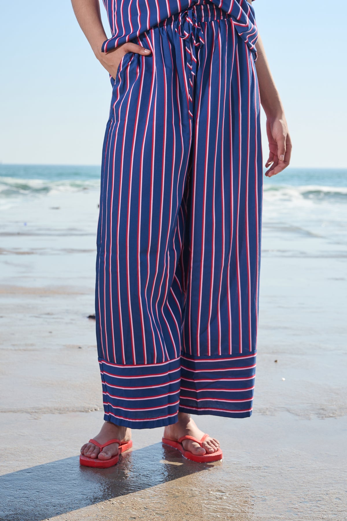 A person standing on a beach wearing navy wide-leg pants with red and white vertical stripes, paired with red flip-flop sandals.