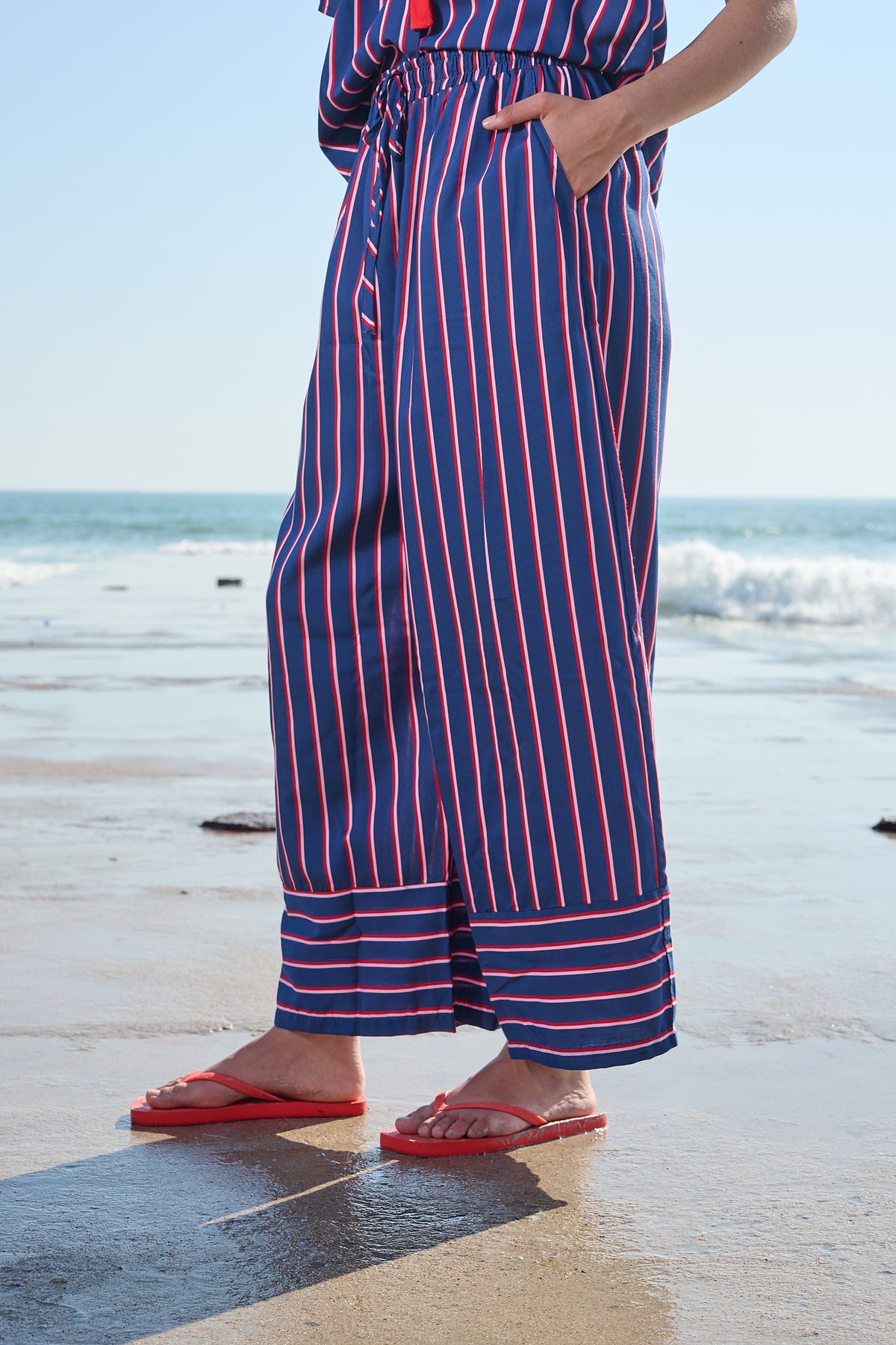A person standing on a beach wearing a blue, red, and white vertically striped wide-leg pant with red flip-flops.