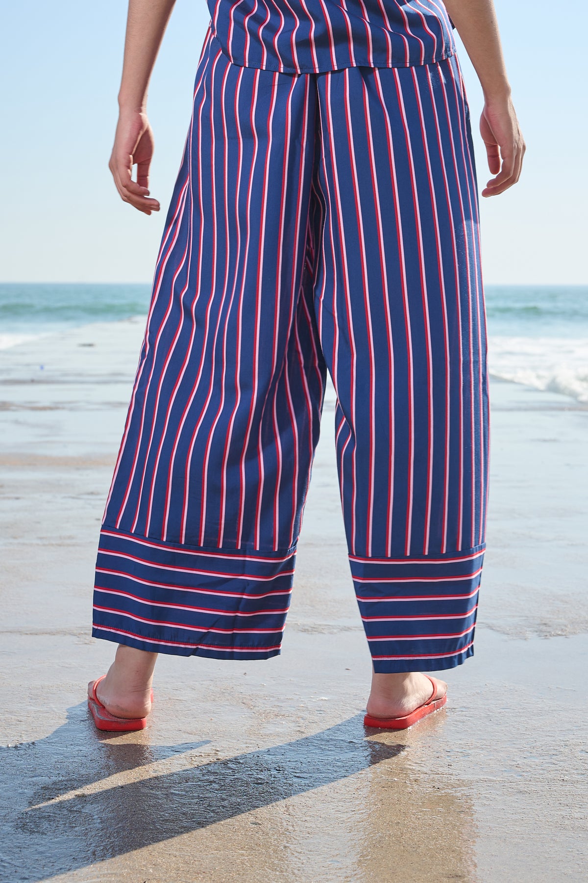 A person standing on a beach wearing loose, wide-leg striped pants in navy blue with red and white vertical stripes, paired with red sandals.