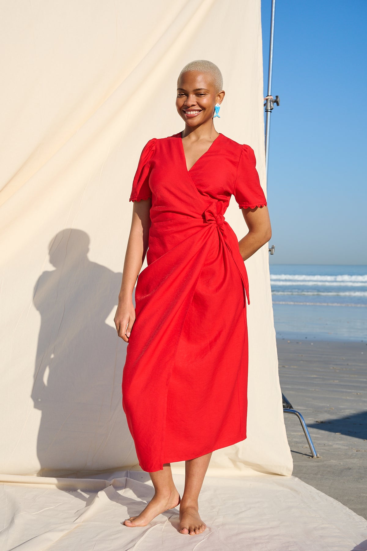 A woman stands barefoot on a beach in front of a large cream fabric backdrop, wearing a bright red wrap-style midi dress with short sleeves and a side tie detail.