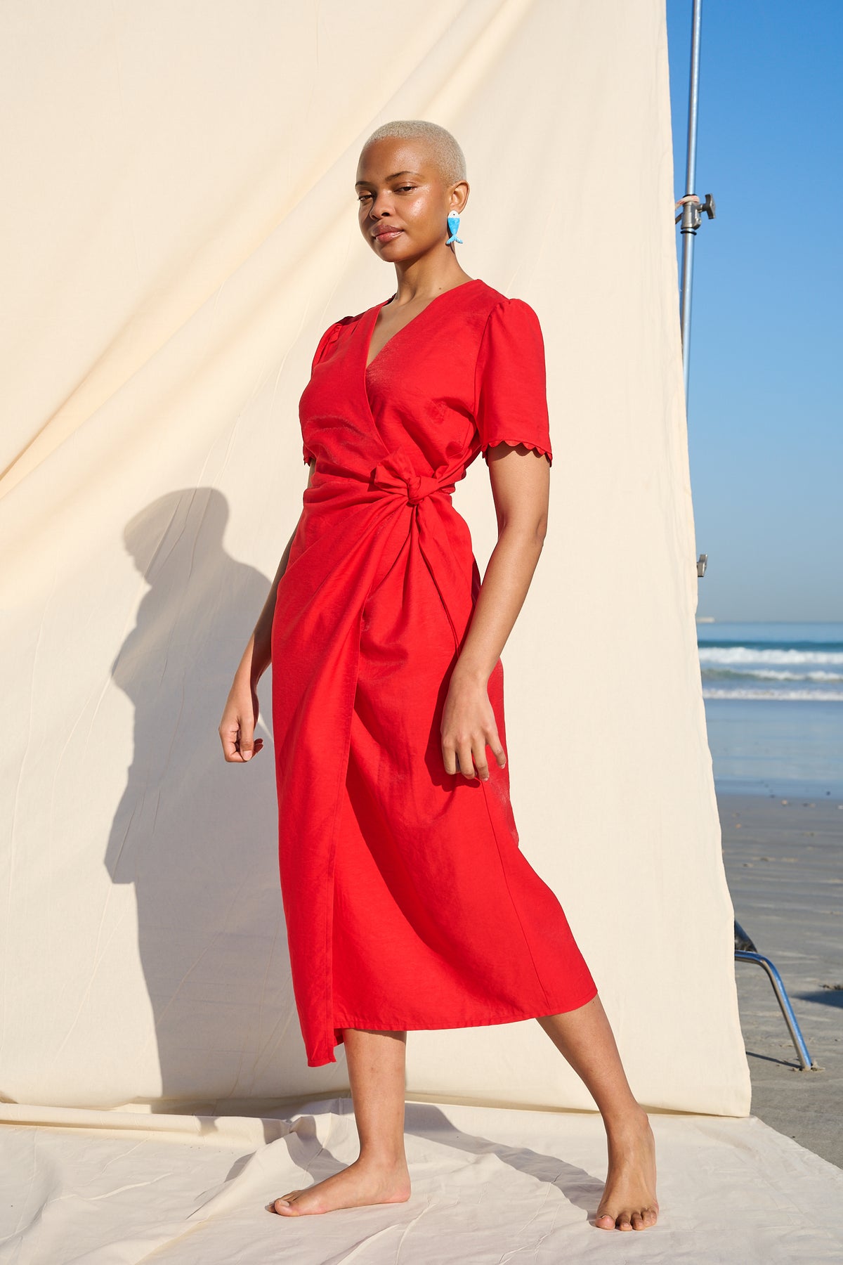 A woman wears a bright red midi wrap dress with short sleeves and a tied waist, standing barefoot on a beach set with a large cream backdrop and ocean visible in the background.