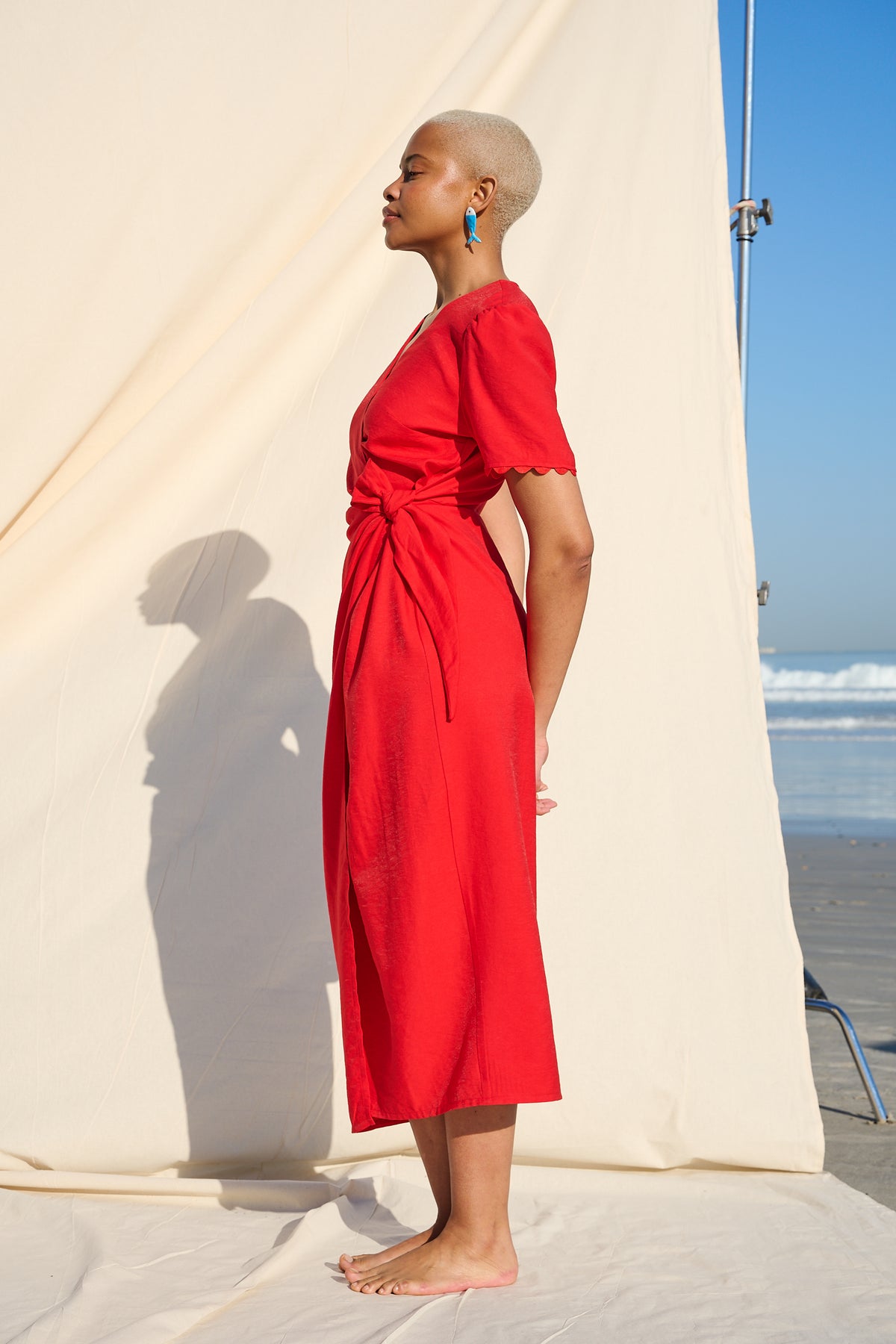 A woman stands in profile outdoors wearing a bright red midi dress with short sleeves and a wrap waist detail. She is barefoot on a white fabric backdrop with the ocean and blue sky visible behind her.