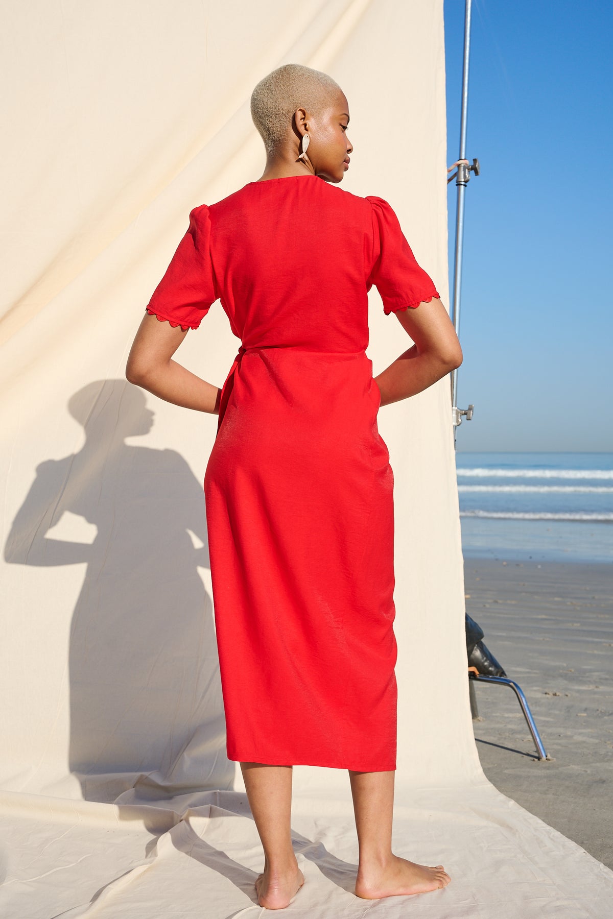 A model stands barefoot on a beach, wearing a bright red midi dress with short sleeves and a waist tie, photographed from behind against a cream backdrop and blue sky.