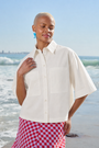 A woman stands on a beach wearing a short-sleeve white button-up shirt with two chest pockets and a red-and-white gingham skirt.