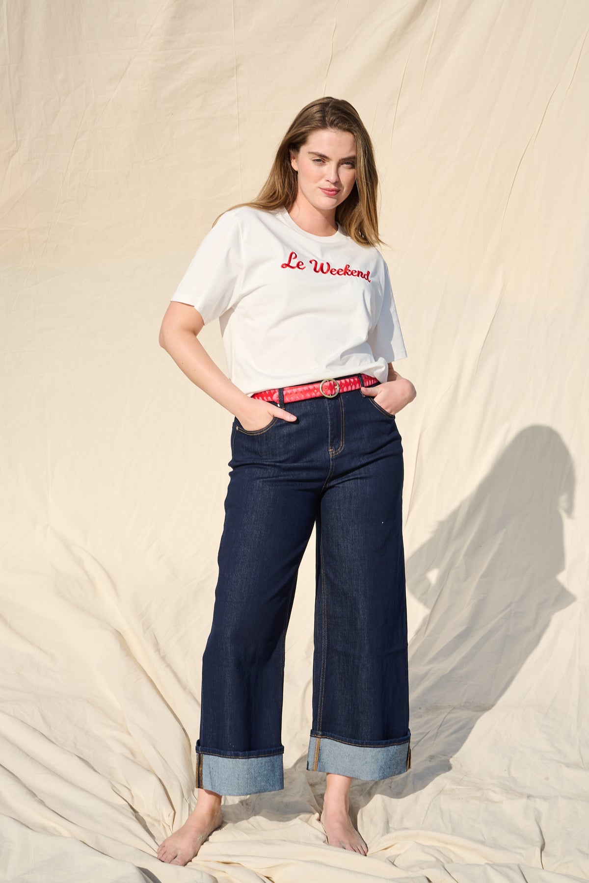 A woman stands barefoot against a draped beige fabric backdrop, wearing a white graphic T-shirt with red embroidered script text reading 'Le Weekend,' dark blue wide-leg jeans with turn up cuffs, and a red leather belt.