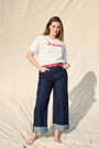 A woman stands barefoot against a draped beige fabric backdrop, wearing a white graphic T-shirt with red embroidered script text reading 'Le Weekend,' dark blue wide-leg jeans with turn up cuffs, and a red leather belt.