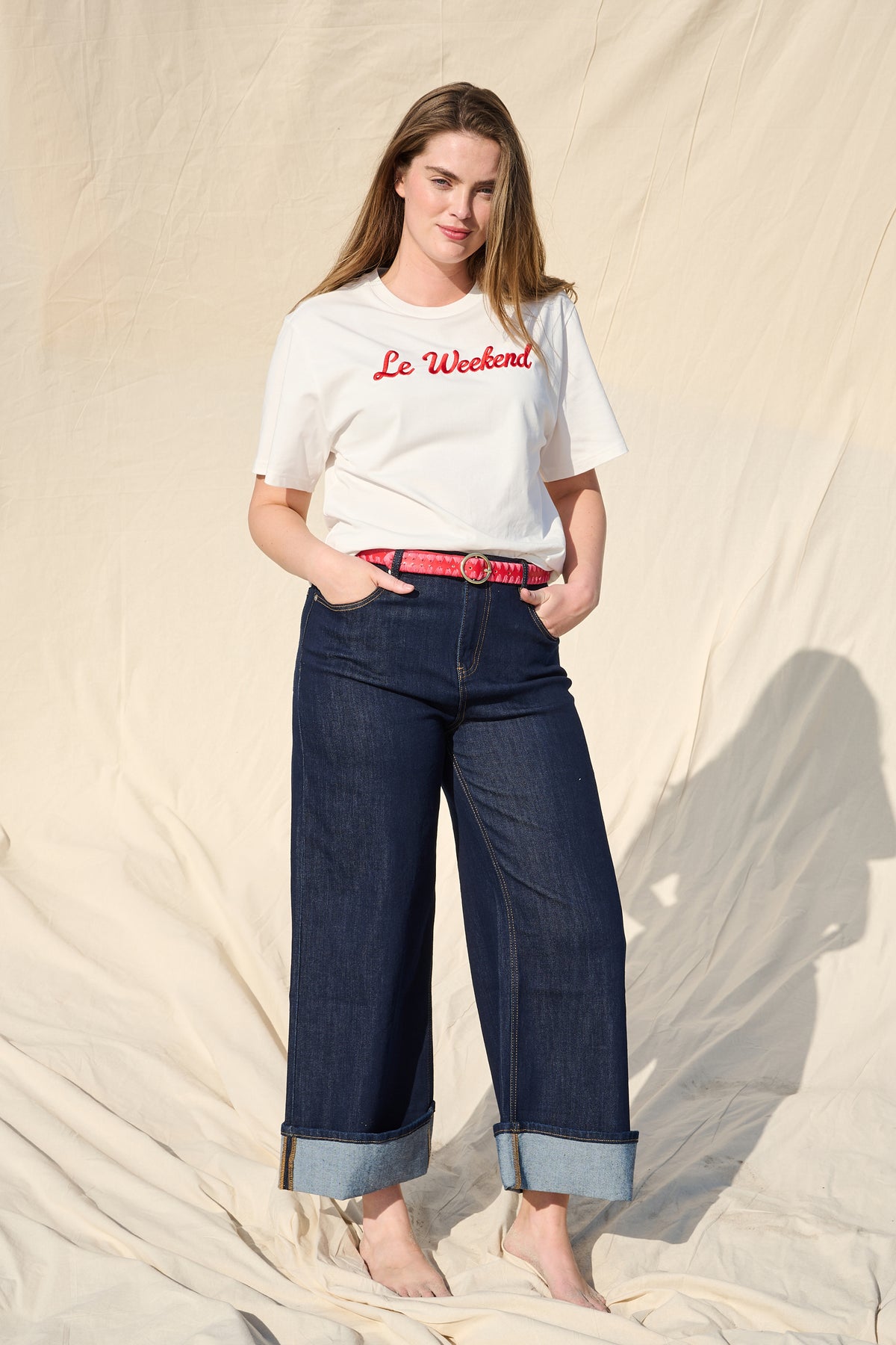 A woman stands barefoot against a draped beige fabric backdrop, wearing a white short-sleeve T-shirt with red embroidery 'Le Weekend,' dark wide-leg jeans with rolled cuffs, and a red patterned belt.