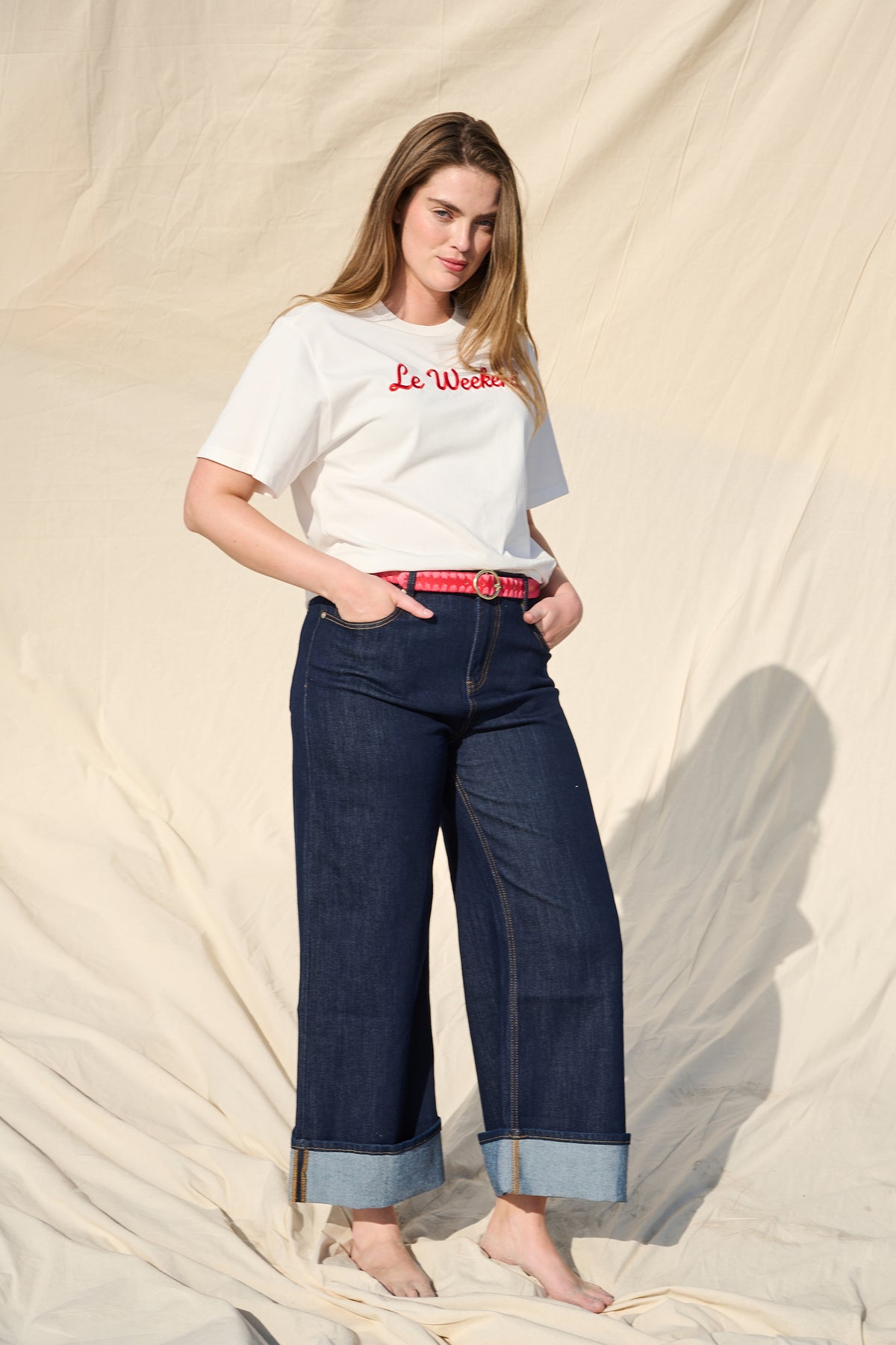 A woman modeling a white crewneck t-shirt with red 'Le Week' embroidery, paired with dark blue wide-leg jeans and a red patterned belt, standing against a draped beige fabric backdrop.