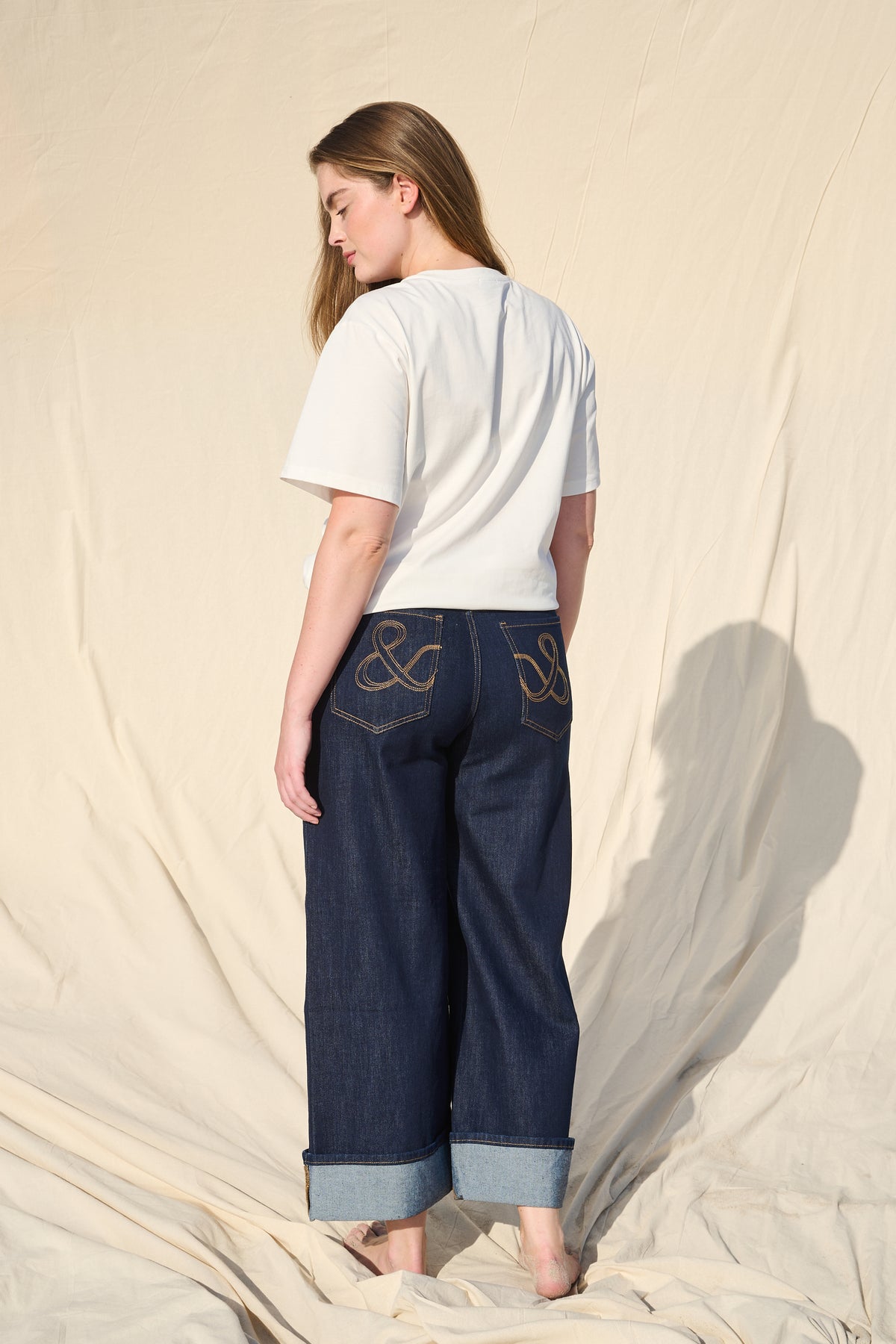 A woman stands barefoot against a draped beige fabric backdrop, shown from the back. She is wearing a white short-sleeve top and dark blue wide-leg cropped jeans with rolled cuffs and gold embroidery on the back pockets.