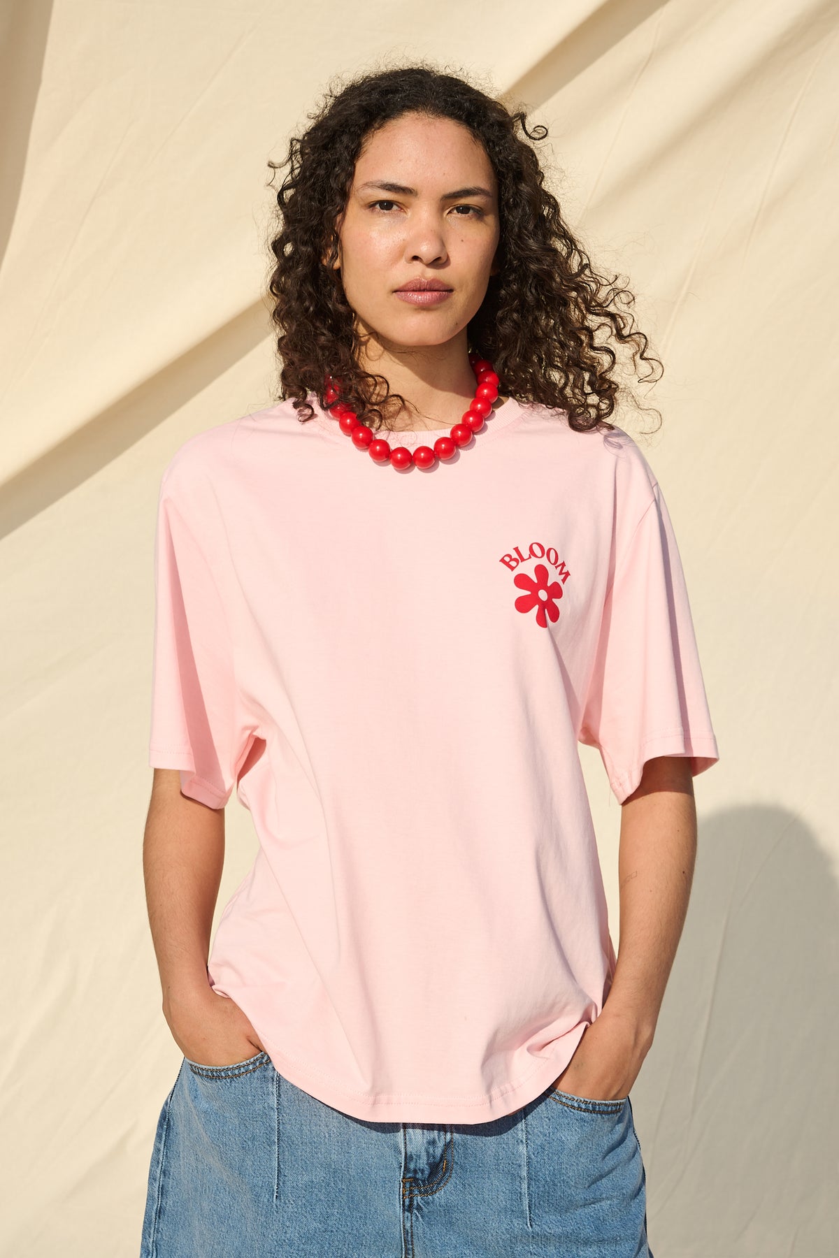 A model with curly dark hair wearing a light pink short-sleeve t-shirt with a red 'BLOOM' flower detail on the left chest, paired with blue denim shorts and a red beaded necklace, standing against a beige fabric backdrop.