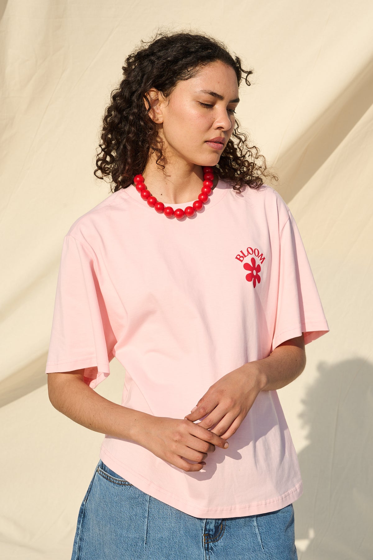 A woman wearing a light pink short-sleeve t-shirt with a red floral graphic and 'BLOOM' text on the chest, paired with blue shorts  and a red beaded necklace, set against a draped beige fabric backdrop.