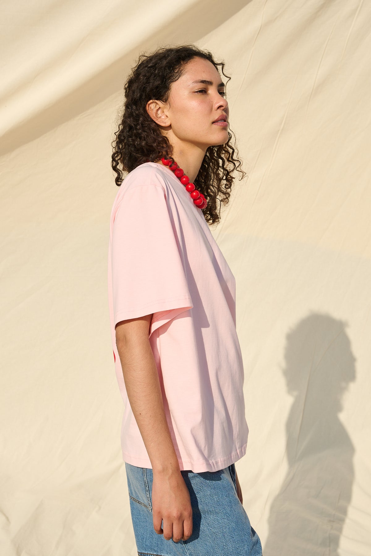 A woman with curly dark hair is shown in profile wearing a light pink short-sleeve top and blue denim shorts, standing against a draped cream fabric backdrop.