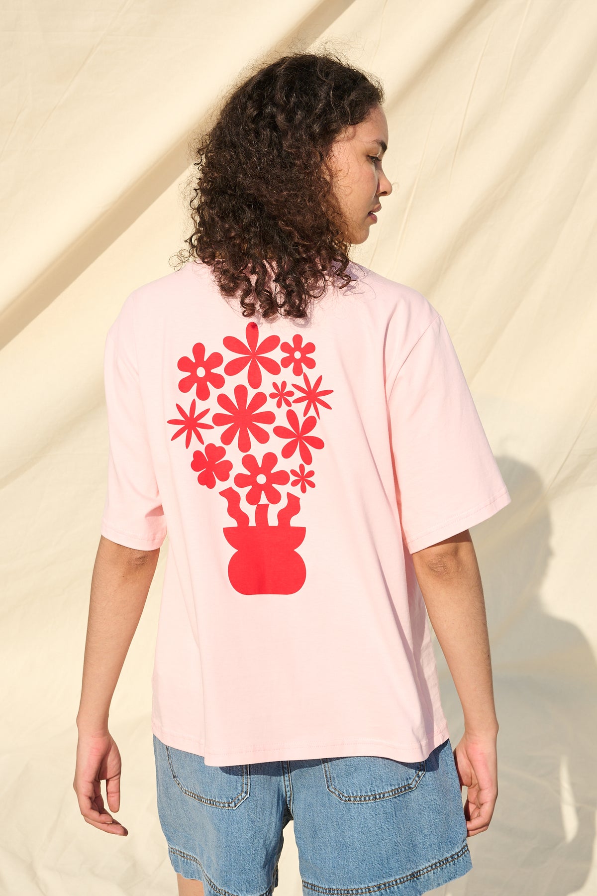 A model wearing a light pink t-shirt with a red graphic of flowers in a pot, shown from the back against a beige fabric backdrop.