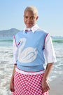 A model stands on a beach wearing a light blue knit sweater vest with a white swan graphic and red trim, layered over a white collared shirt and paired with a red and white gingham skirt.