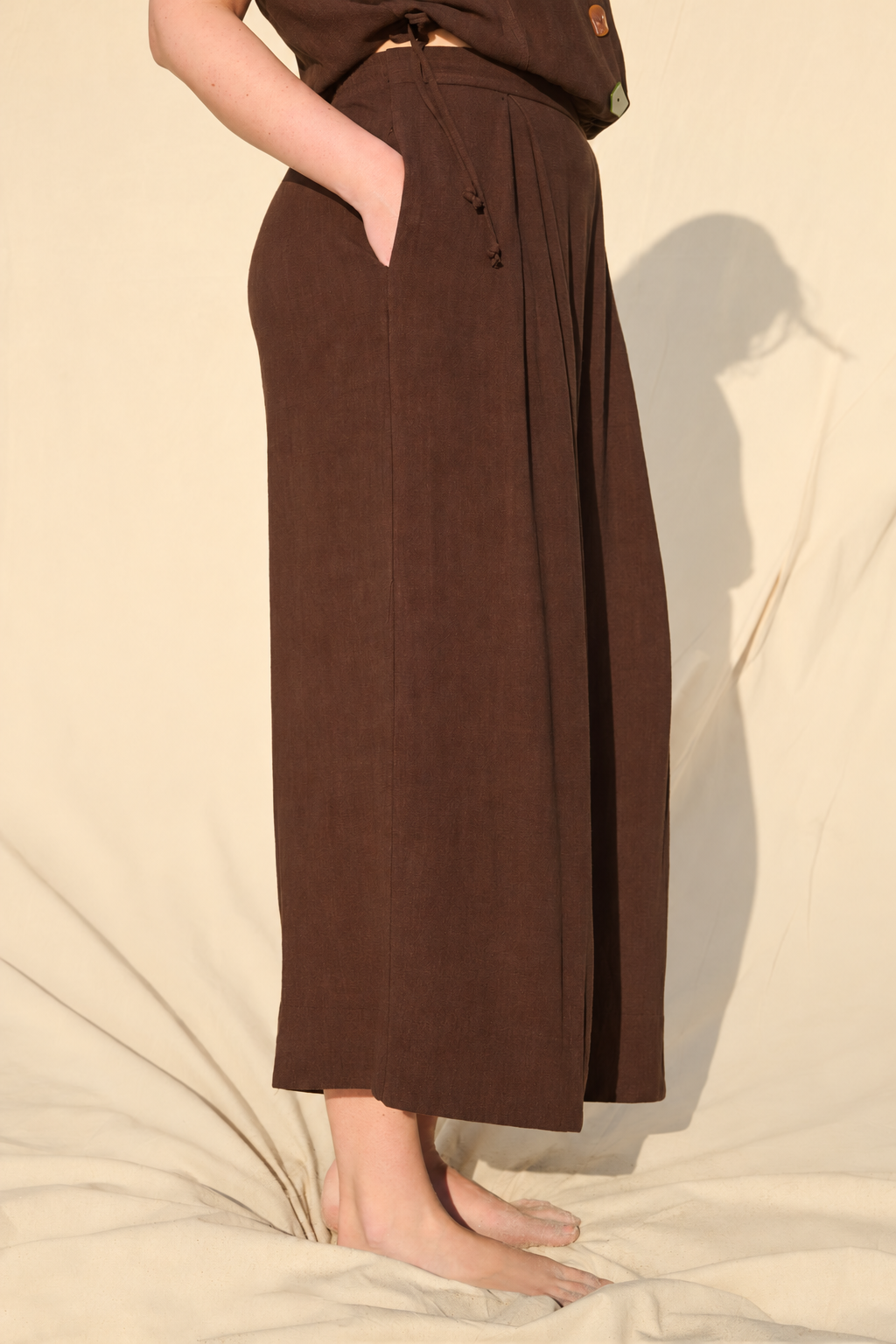 A person wearing dark brown wide-leg cropped pants with side pockets, standing barefoot against a warm beige fabric backdrop.