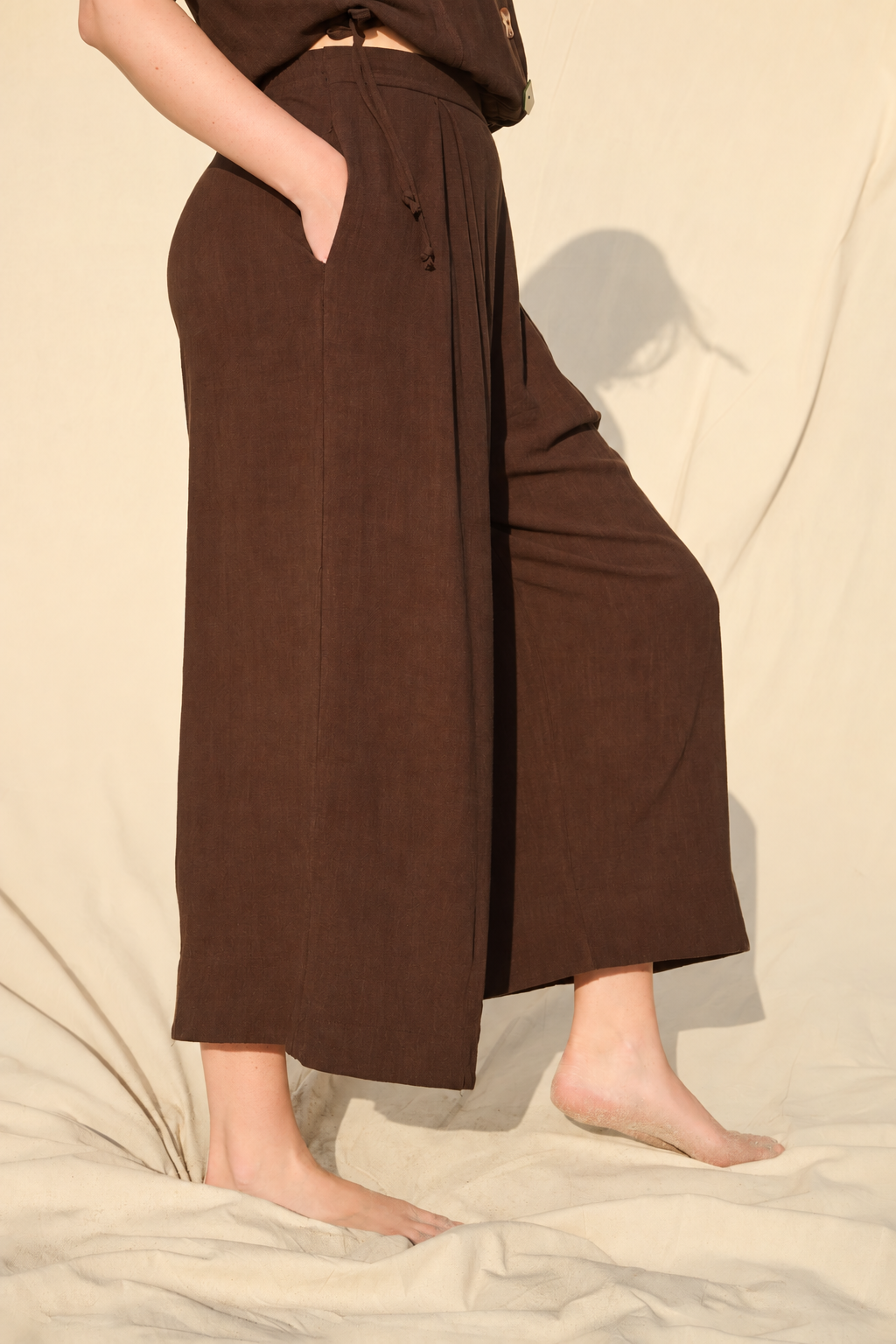 A person wearing dark brown wide-leg cropped pants with side pockets, standing barefoot on a beige fabric backdrop.