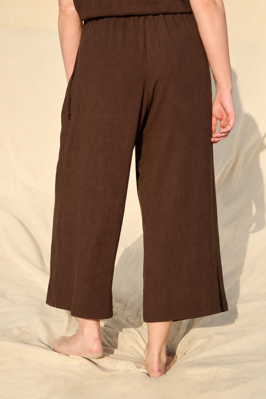 A person wearing loose, wide-leg cropped pants in a dark brown color, standing barefoot on a draped beige fabric backdrop.