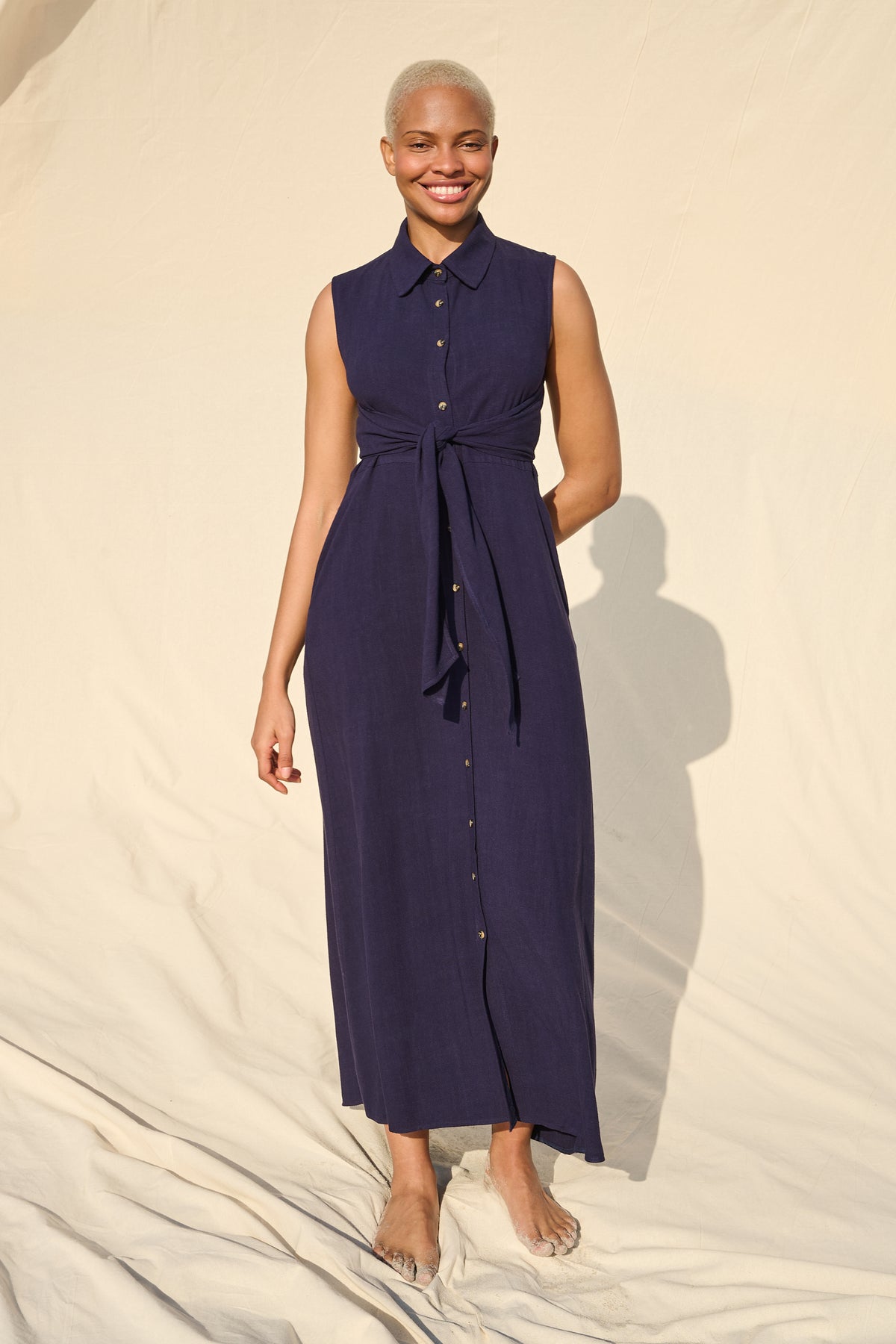 A woman modeling a sleeveless, button-front midi dress in deep navy blue with a matching tie waist, standing against a draped cream fabric backdrop.