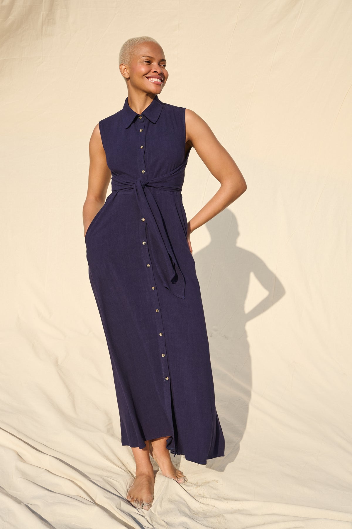 A woman modeling a sleeveless navy blue midi dress with a button-down front, collar, and self-tie waist, standing against a draped cream fabric backdrop.