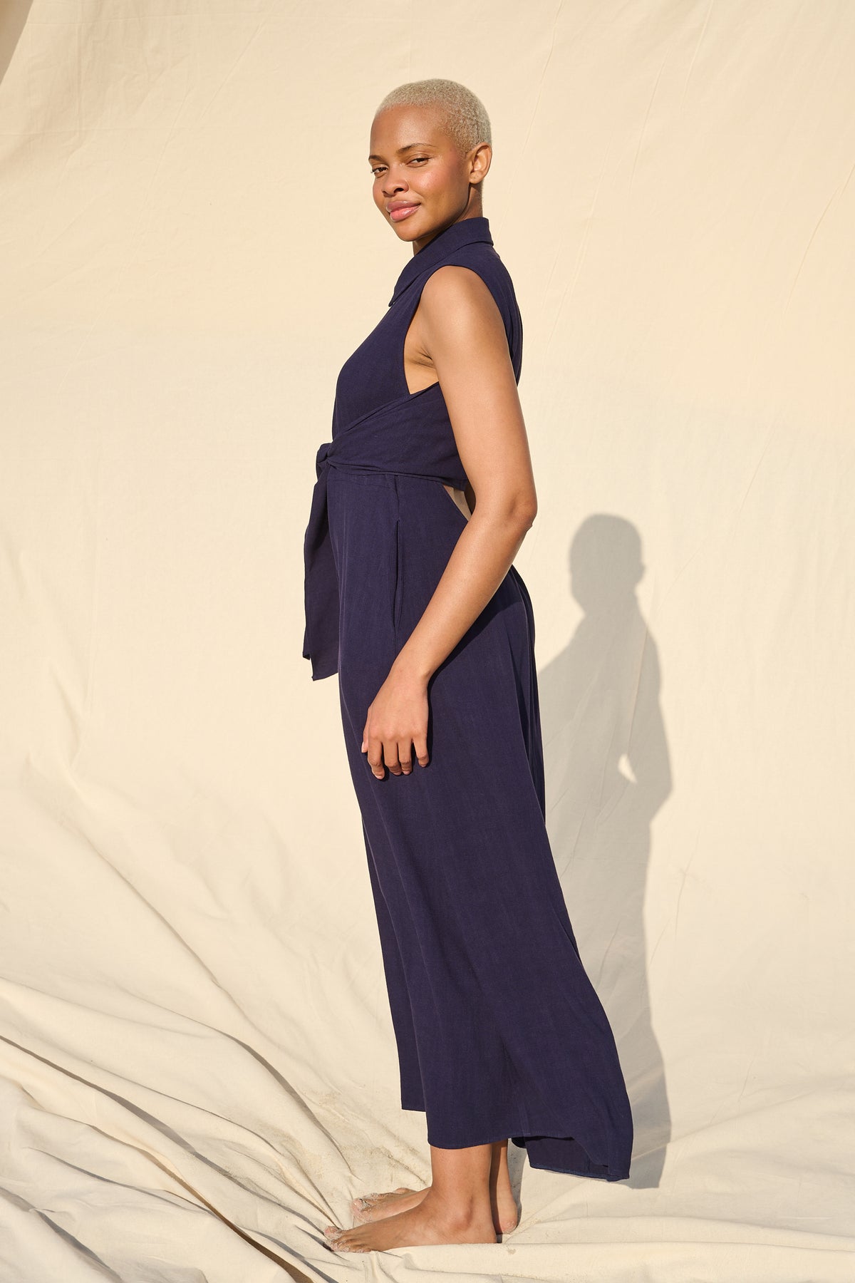 A model stands in profile wearing a sleeveless navy blue maxi dress with a high collar and a tied waist sash, posed against a draped cream fabric backdrop with a distinct shadow.