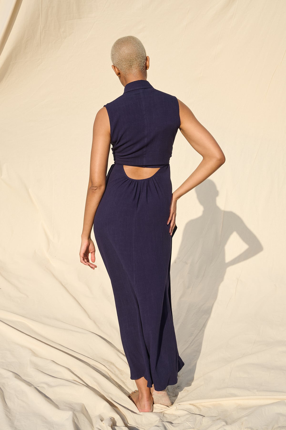 A model from behind wearing a sleeveless navy blue maxi dress with a high mock neck and a cutout at the waist, standing against a draped beige fabric backdrop.