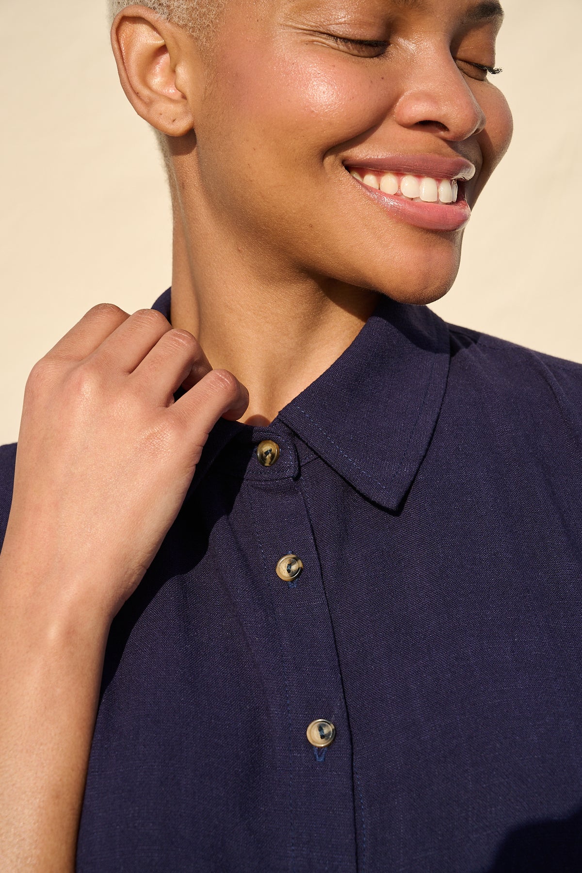 Close-up of a smiling person wearing a dark navy button-front collared shirt, with warm sunlight and a plain beige background.