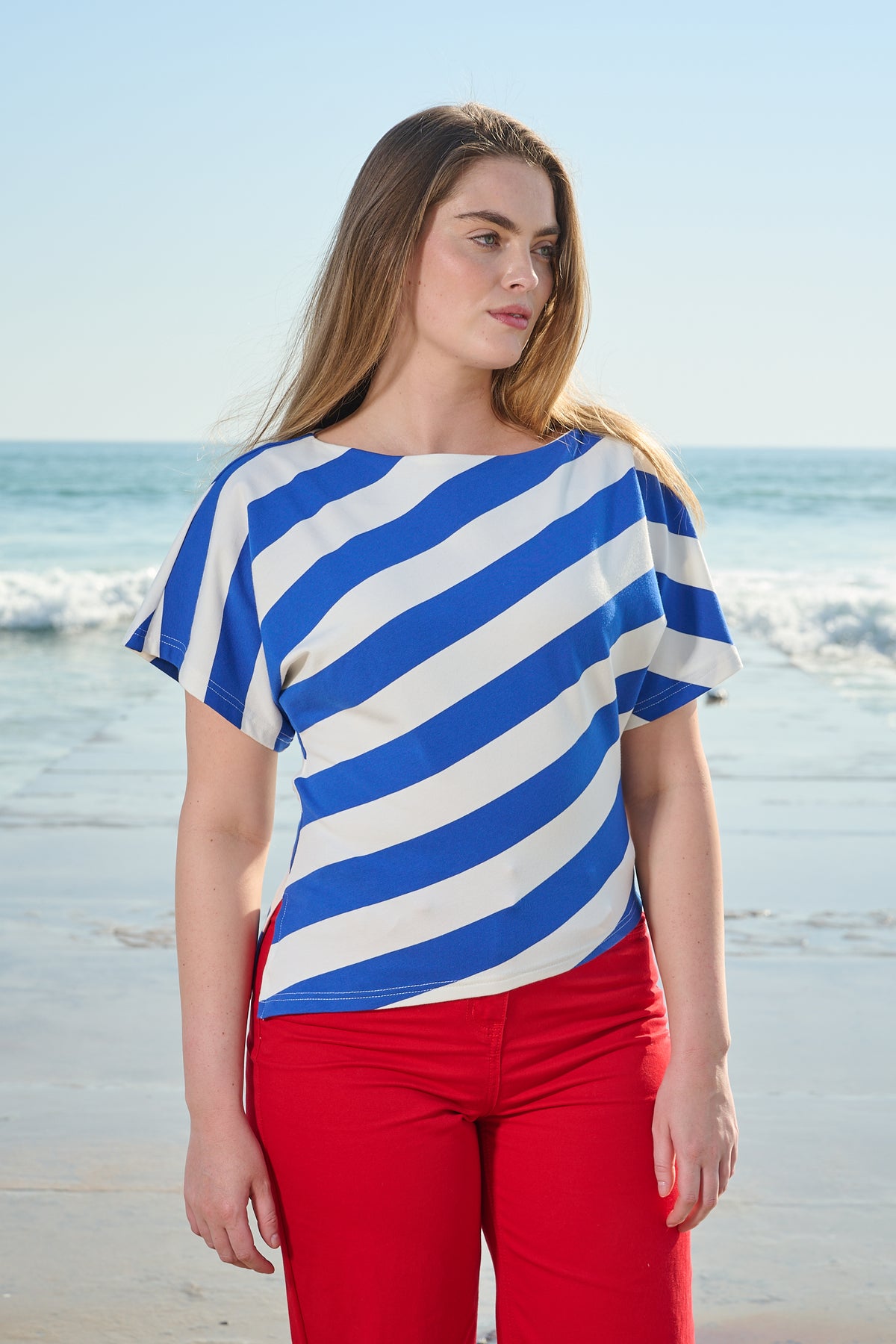 A woman standing on a beach wearing a blue and white diagonally striped short-sleeve top and bright red pants.